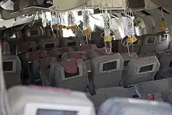 Interior of the aircraft, after the crash, showing oxygen masks hanging from the ceiling, ready for use. The seat backs have video displays installed, and most seats are in position, some reclined, some upright. The overhead compartments are open.