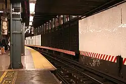 View of the southbound local platform of the 14th Street–Union Square station on the IRT Lexington Avenue Line. Behind the wall and the black bars on the right there is an abandoned side platform, whose edge is still visible. The mezzanine is right above. The red and white stripe indicates that there is no clearance between trains and the wall. In the foreground one can see the gap fillers that are automatically operated via proximity sensors. The writing on the floor says "stand clear".