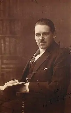 A man with rimless glasses, slicked back hair, and a toothbrush mustache in an ill-fitting suit sitting in a chair and holding an open book