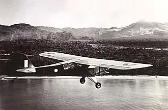 Single-engined high-wing light plane flying over water, with jungle terrain in background