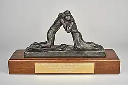 Photograph of The Coventry International Prize for Peace and Reconciliation awarded to Dr Madeleine Sharp in 2004. The award is a metal sculpture of two figures embracing, mounted onto a wooden block with a gold-coloured engraved plaque on the front bearing the inscription "The Coventry International Prize for Peace and Reconciliation Awarded to Dr Madeleine Sharp 14th November 2004 Father Forgive".