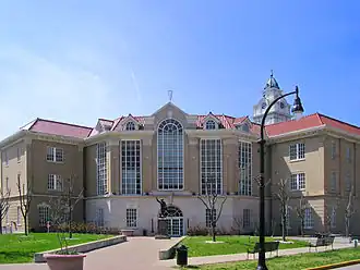 A large, brown building with towering windows, a clock tower, and a statue of a man in front