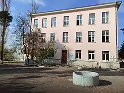 A three-story, rectangular, pale pink building with many windows. In the foreground is a dirt yard with a small tree and a circular concrete well structure.