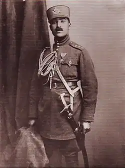 Sepia photograph of Mahmud Khan Pulādeen standing in a military uniform, there is a cloth on the left-hand side of the wall behind him