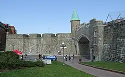 Quebec's restored city wall is gray stone about 20 feet (6.1 meters) high. The St. Jean's gate has a modern road going through it, and has a copper-roofed turret on the left bastion. A paved path goes through a grassy area below the wall.