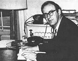 A middle-aged man in a suit, wearing glasses and seated at a desk, looking to the side at the camera.