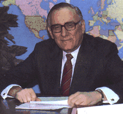 Middle-aged man with black and gray hair sitting behind a desk, wearing a navy blue suitcoat, white dress shirt, red necktie, and black-rimmed eyeglasses