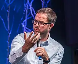 A man with a beard and glasses gestures while holding a microphone.