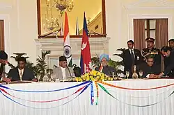 Mr. Sharat Singh Bhandari is shown at a formal table in front of Nepalese flags in his capacity as Minister of Tourism and Civil Aviation signing an air and railway infrastructure agreement with the Union Minister For External Affairs, Shri S.M. Krishna. A large mirror adorns the wall over an ornate mantelpiece in the background. An unknown person in a smart suit is also in attendance, standing in the right background of the photo.
