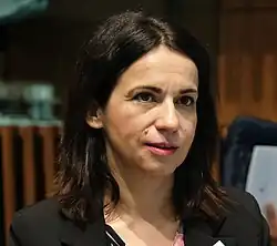Silvia Calzón, wearing a light turquoise jacket over a black top, sits and looks slightly to the side in front of a black microphone and a pump-bottle of hand sanitizer.