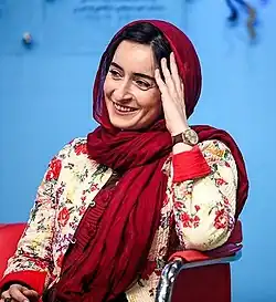 35jährige Frau auf Stuhl sitzend, freundlich lächelnd, mit Kopftuch, den Kopf leicht mit dem li. Arm abstützend