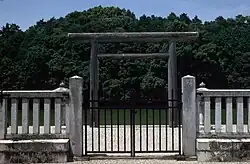Torii gate behind a concrete fence. In the back behind the torii there is water and behind it trees.