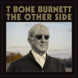 T Bone Burnett in black and white wearing sunglasses in front of a field backdrop in color