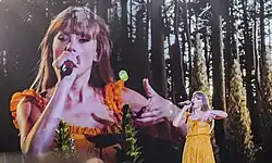 Swift singing onto a mic as a background screen shows her; she is wearing an orange dress.