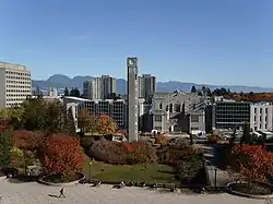 Photograph of a clock tower, standing in a park framed in autumn coloured trees. Beyond the tower are six high-rise grey buildings and behind those are mountain peaks.