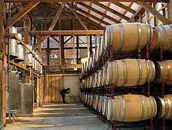 A wood-paneled warehouse with stainless steel fermentation tanks on the left, and rows of oak barrels on the right.