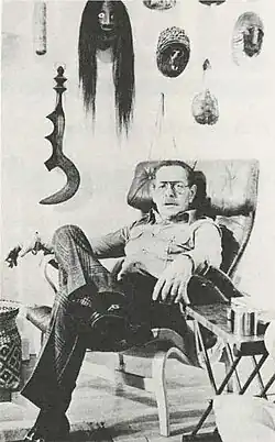 A white man with glasses seated with his legs crossed in front of a wall adorned with various masks and objects