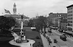 An urban street and park with a large Romanesque building. Two streetcars are on the street.